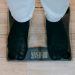 From above of crop unrecognizable plus size person in casual clothes standing on weighing scale on wooden floor