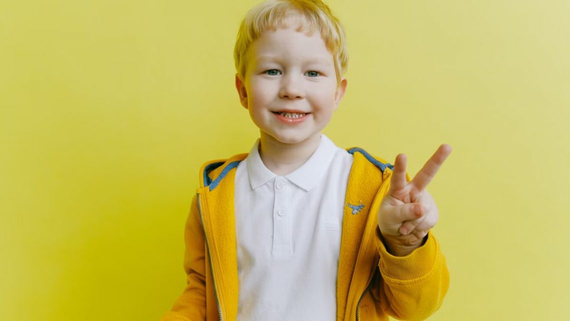 Smiling boy making peace sign in a yellow jacket against a bright background.