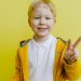 Smiling boy making peace sign in a yellow jacket against a bright background.